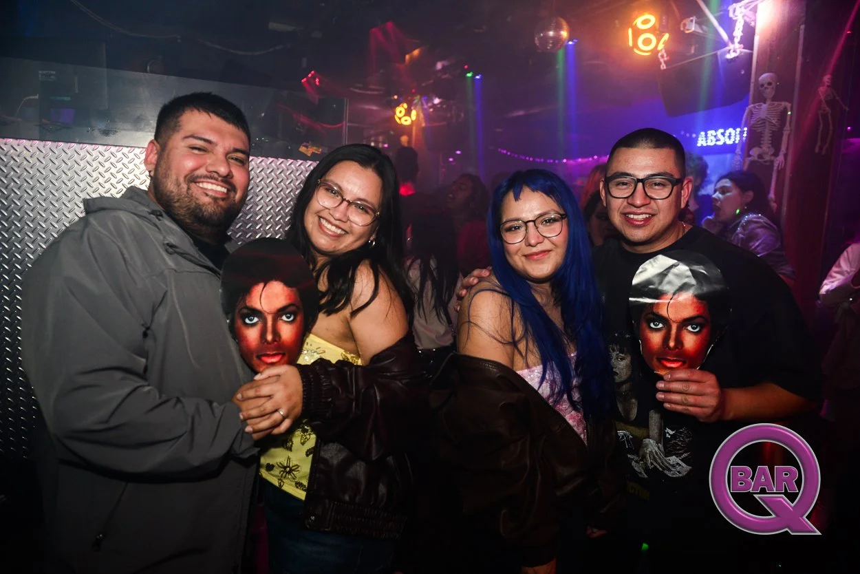 Four friends in a nightclub, smiling and holding images of Michael Jackson, with colorful lighting, skeleton decorations, and a sign that says 'ABSOLUT' in the background.