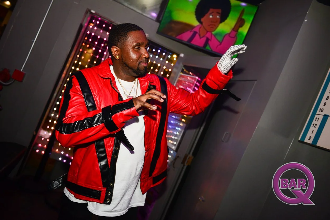 A man in a red and black leather jacket with a white shirt underneath is dancing at a bar. He is wearing white gloves with small jewels. Behind him, there are colorful lights and a TV screen showing a cartoon of a woman with an afro hairstyle.