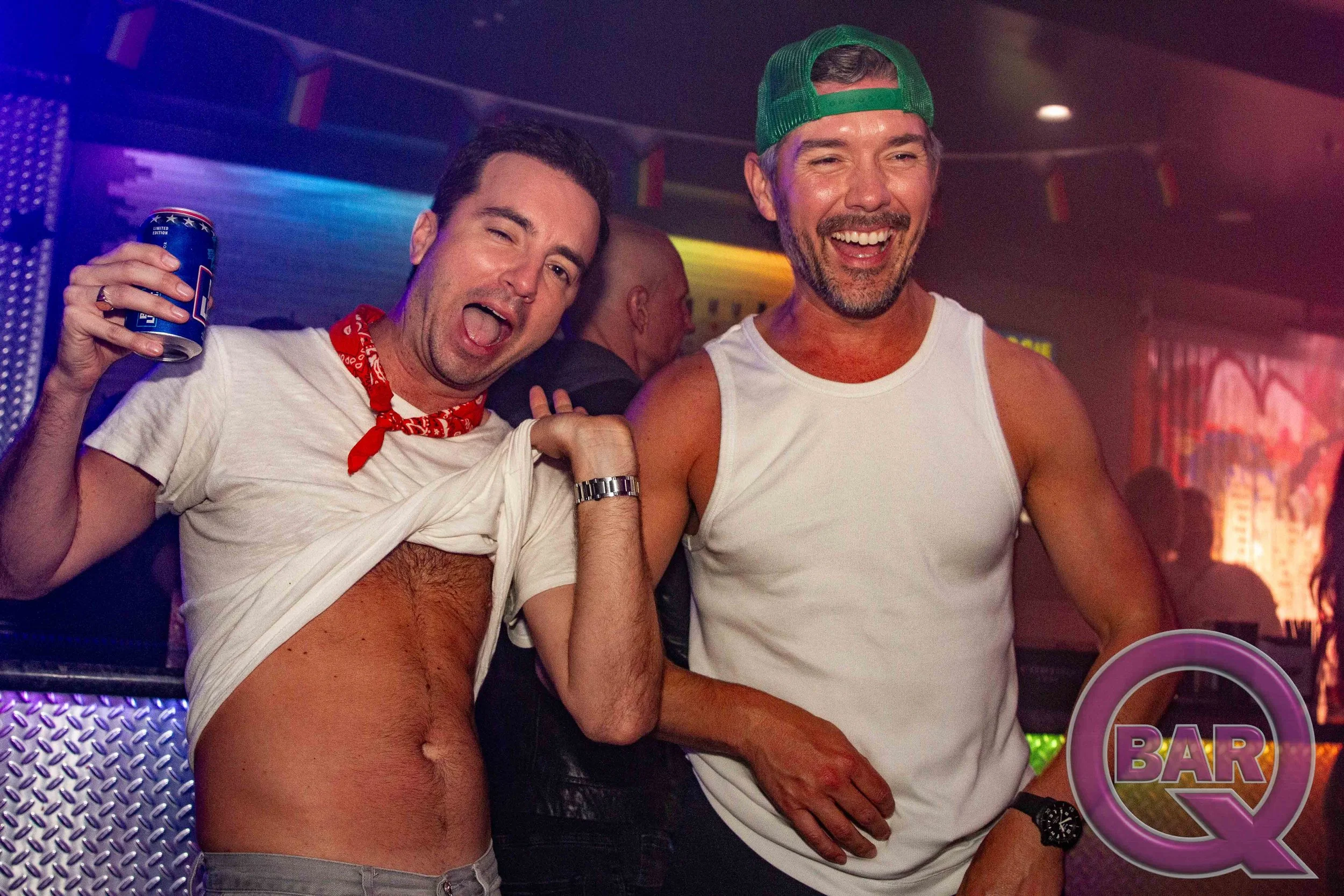 Two men smiling and celebrating in a nightclub, one holding a can of beer and wearing a red bandana, the other wearing a green cap and white tank top, with colorful lights and a bar sign in the background.