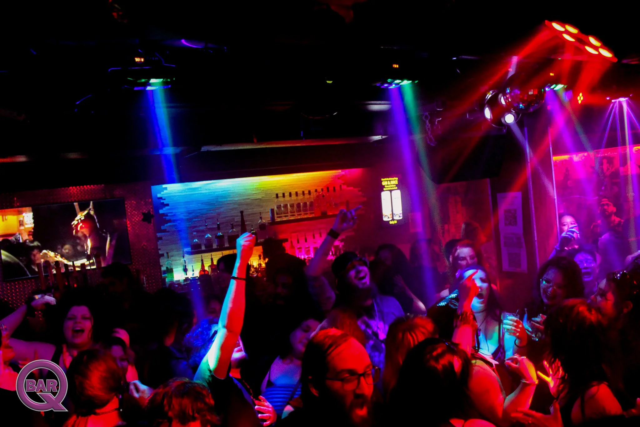 Crowd of people dancing and enjoying a party in a nightclub with vibrant colorful lights and a bar in the background.