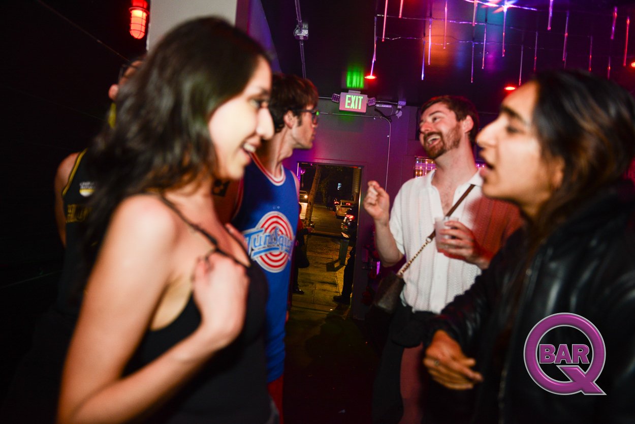 Group of people socializing and laughing inside a nightclub with colorful neon lights and an exit sign in the background.
