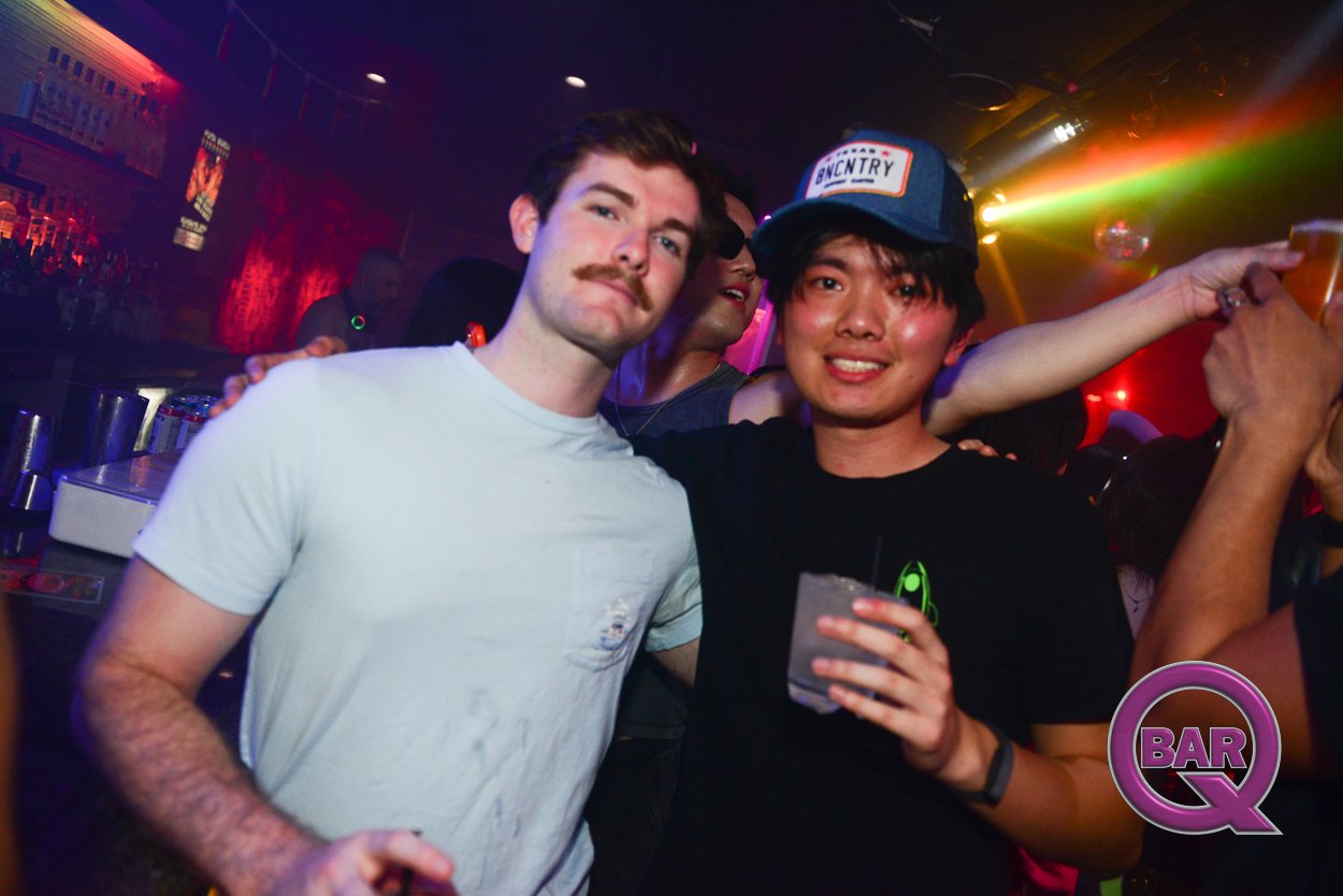 People dancing and having fun at a bar or nightclub with colorful lights, one person holding a drink, and a 'Bar Q' logo in the corner.