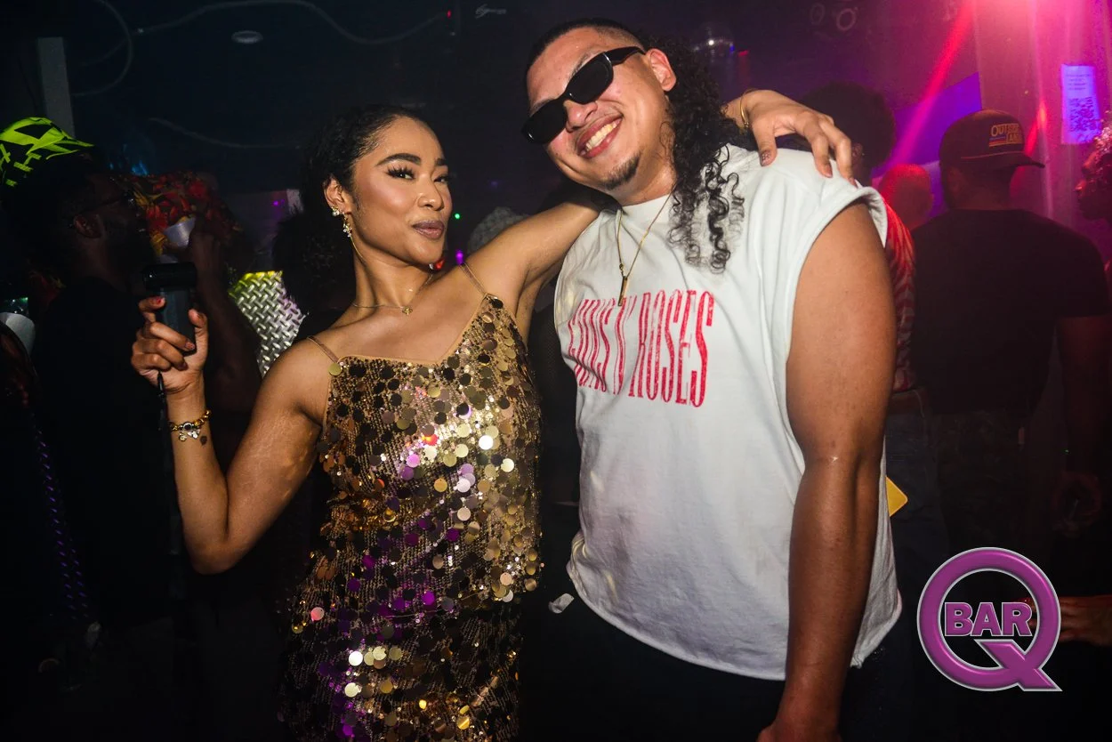 Two people at a nightclub, one woman wearing a gold sequin dress and a man wearing a white T-shirt with red text, sunglasses, and a gold chain, smiling and posing together.