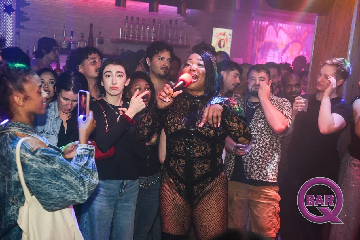 A lively nightclub scene with a diverse group of people watching a female singer perform on stage, holding a microphone, with colorful lighting and a bar in the background.