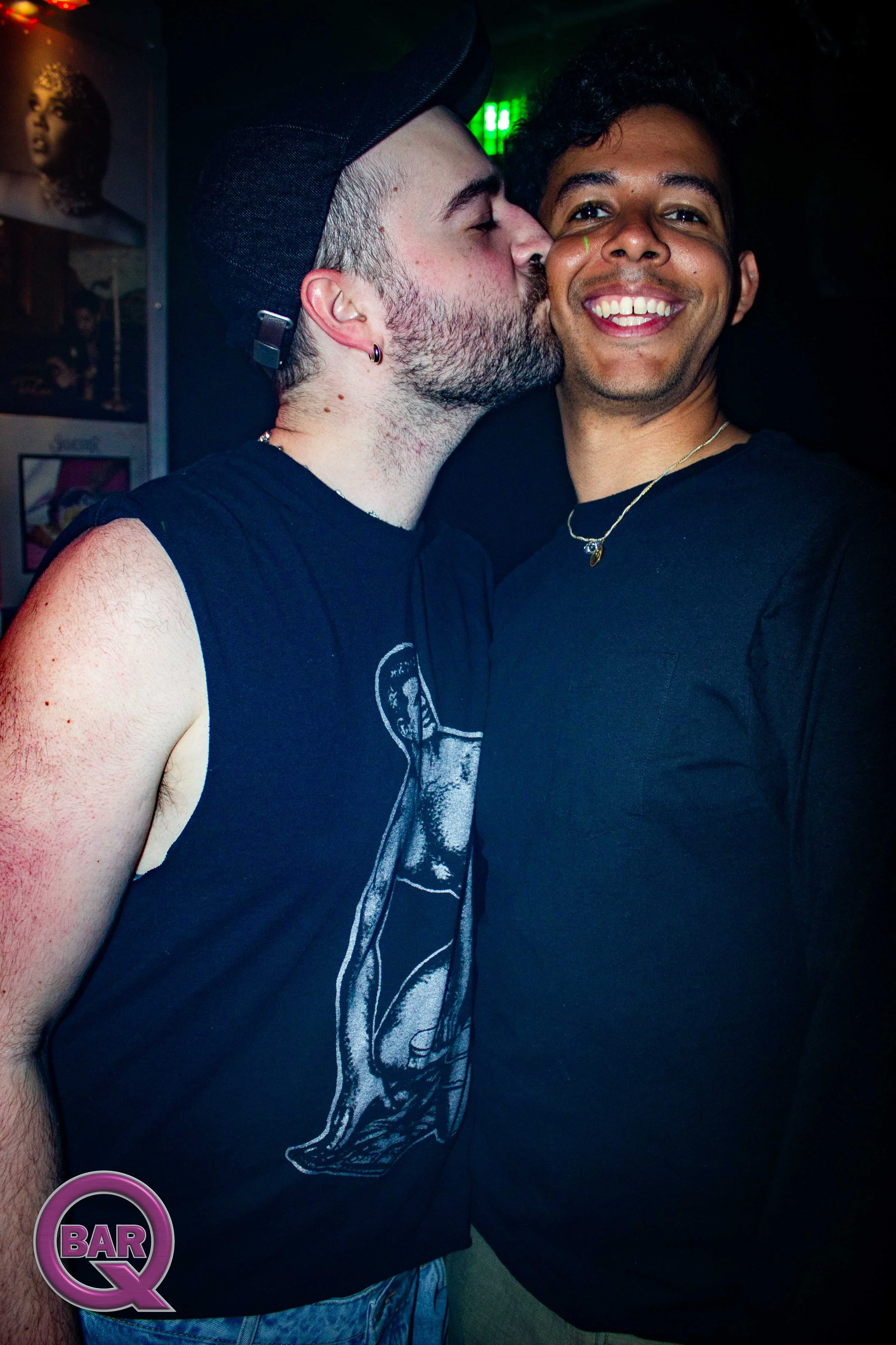 Two men happily pose for a photo, one kissing the other's cheek, both wearing black t-shirts, with a bar logo visible in the bottom left corner.