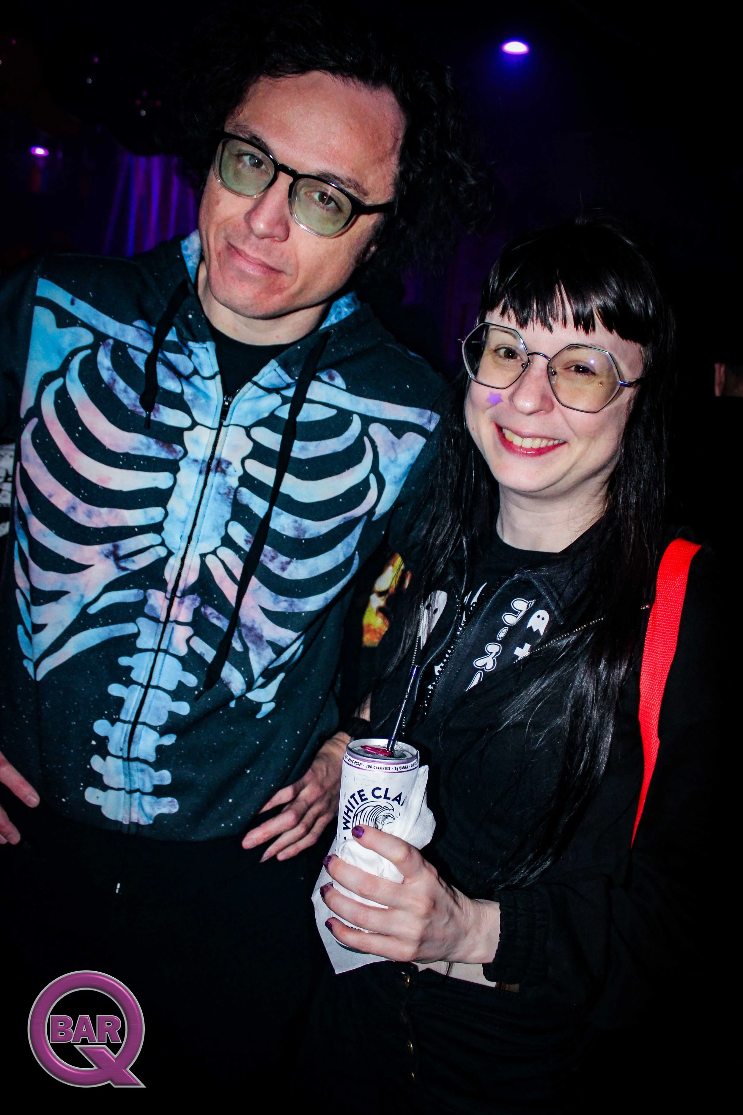 Two people, a man wearing glasses and a skeleton hoodie and a woman with dark hair and glasses, standing together inside a bar or nightclub with purple lighting. The woman is holding a drink in a white paper cup with a straw and a purple star sticker