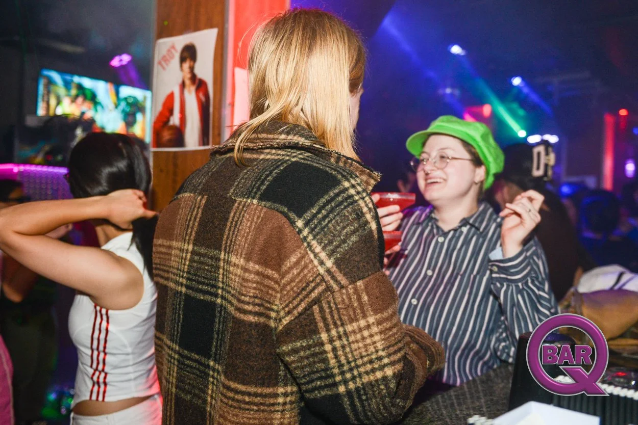 People socializing at a nightclub or bar with colorful lighting, a woman with a striped shirt and green hat smiling, and a poster of a person in a red jacket on the wall.
