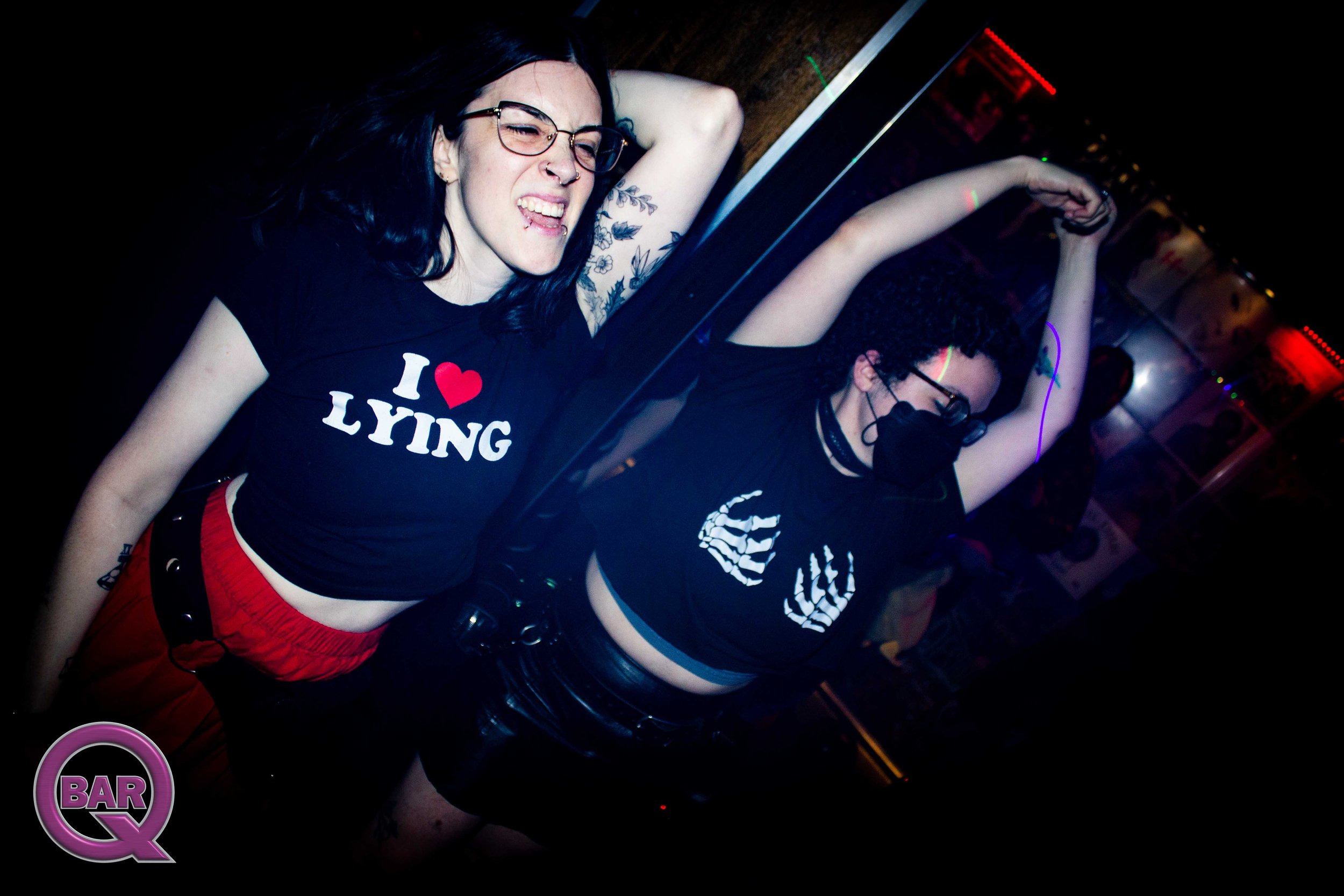 A woman with black hair, glasses, and tattoos, wearing a black shirt with red shorts, dancing with her eyes closed in a bar or club setting with colorful lights.