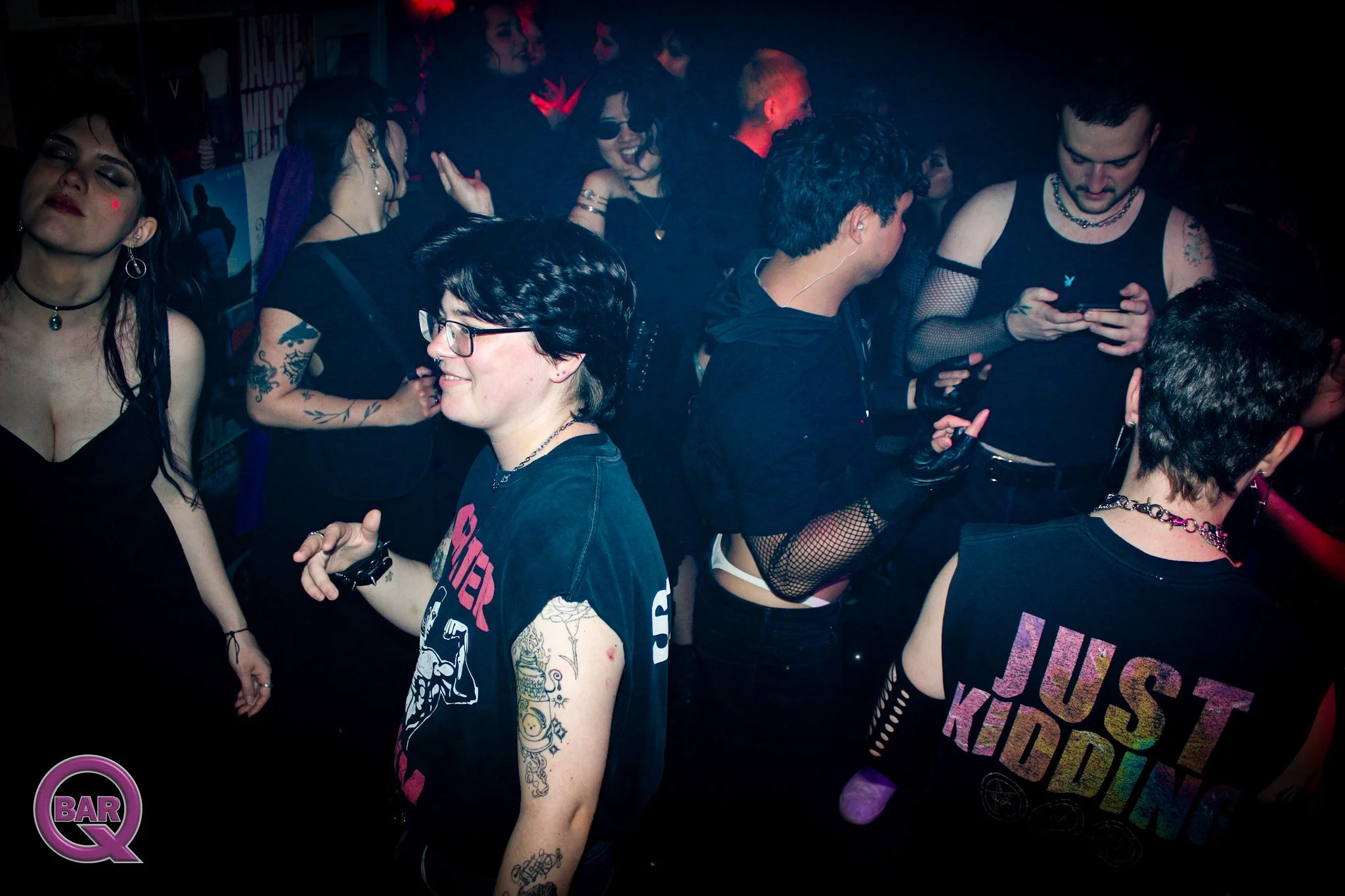 Group of young people at a party or nightclub, some wearing dark clothing, tattoos, and accessories, dancing and socializing in a dimly lit space.