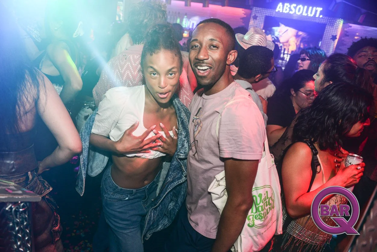 Two people are dancing and smiling at a crowded indoor nightclub with colorful lights, a large illuminated sign that says 'ABSOLUT,' and a neon BAR Q logo in the corner.