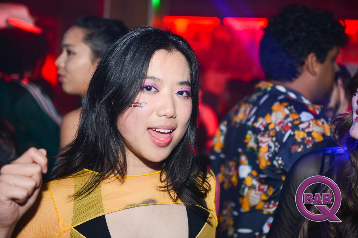 A young woman with black hair and colorful eyeshadow at a party, with a blurred background of other people and colorful lights.