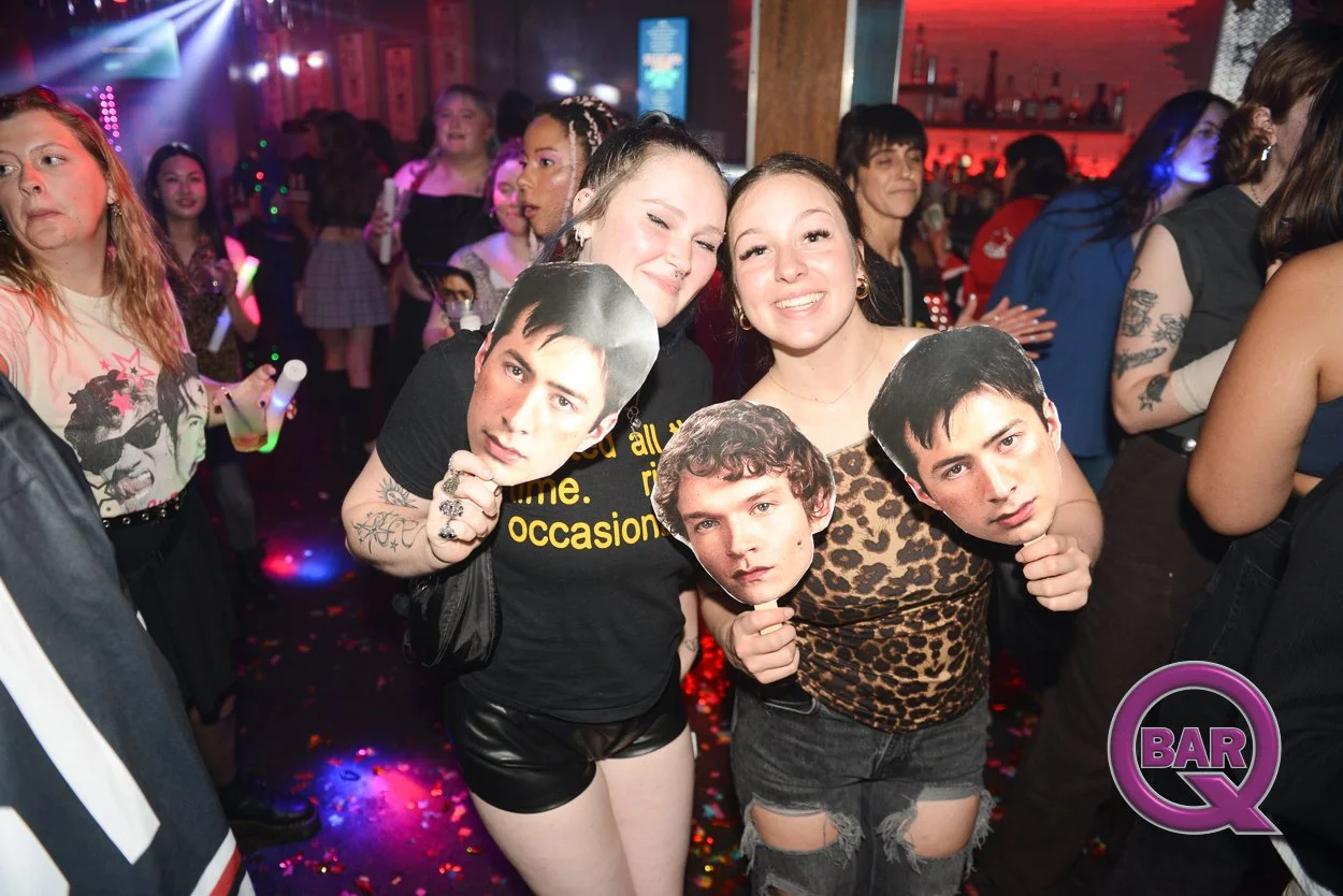 Two women at a bar holding cardboard cutouts of male faces while smiling, with other partygoers in the background, colorful lighting, and a dance floor with confetti.