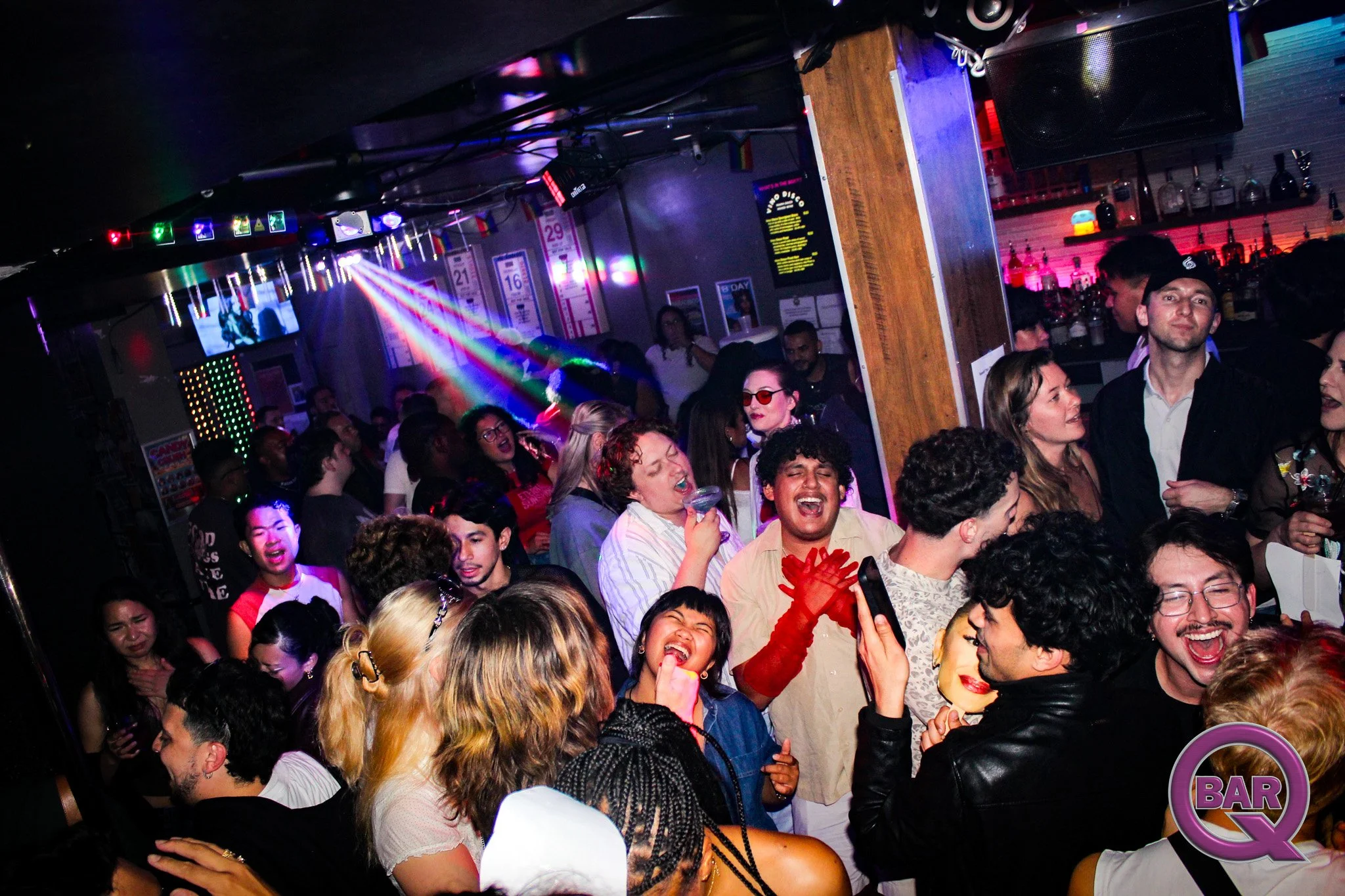 Crowd of people dancing and socializing in a nightclub with colorful lighting, bar area, and a large screen in the background.