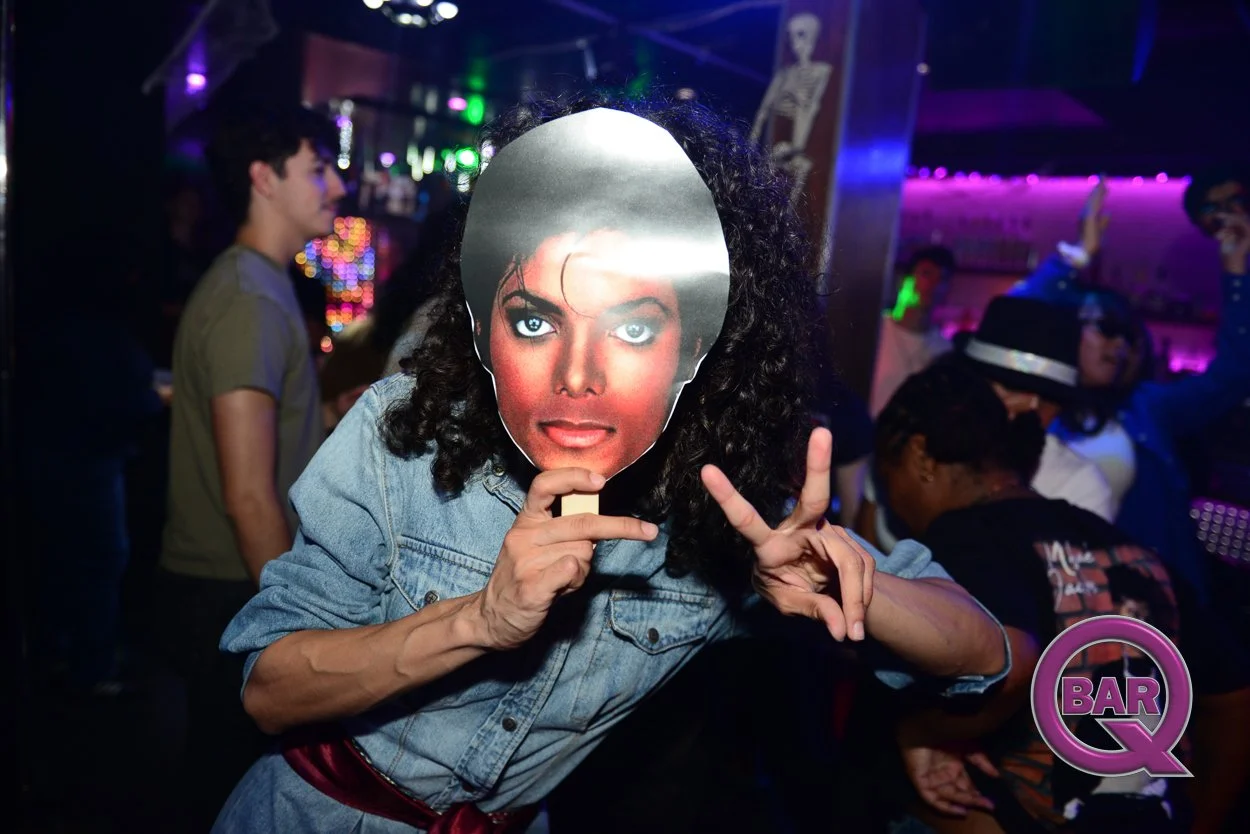 Person holding a large magazine cutout of a woman's face in front of their face while posing with a peace sign at a lively bar or nightclub with colorful lights and other people dancing and socializing.
