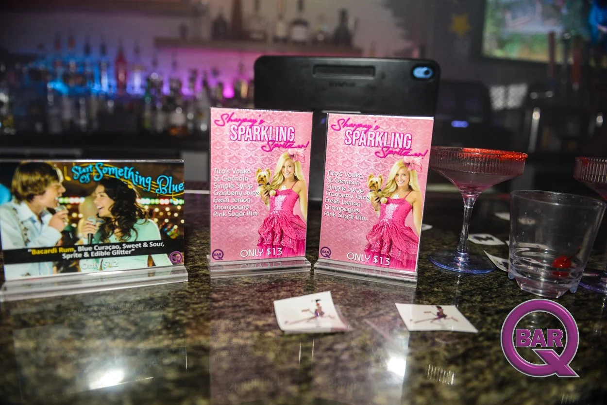 A bar countertop displays promotional cards for drinks; one card features a woman in a pink dress holding a dog with a pink background, advertising a drink for $13, and another card promotes drinks including Tito's vodka, cranberry juice, and champag