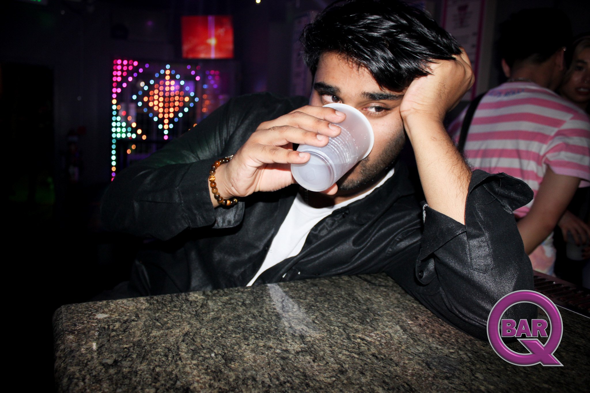A man with dark hair and a beard holding a plastic cup near his face in a bar or nightclub setting, with colorful lights and people in the background.