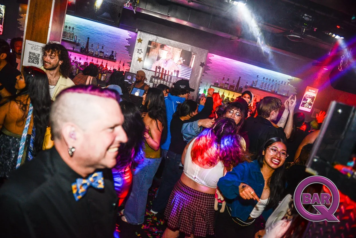 People dancing and enjoying themselves at a lively bar or nightclub with colorful lighting and a neon bar in the background.