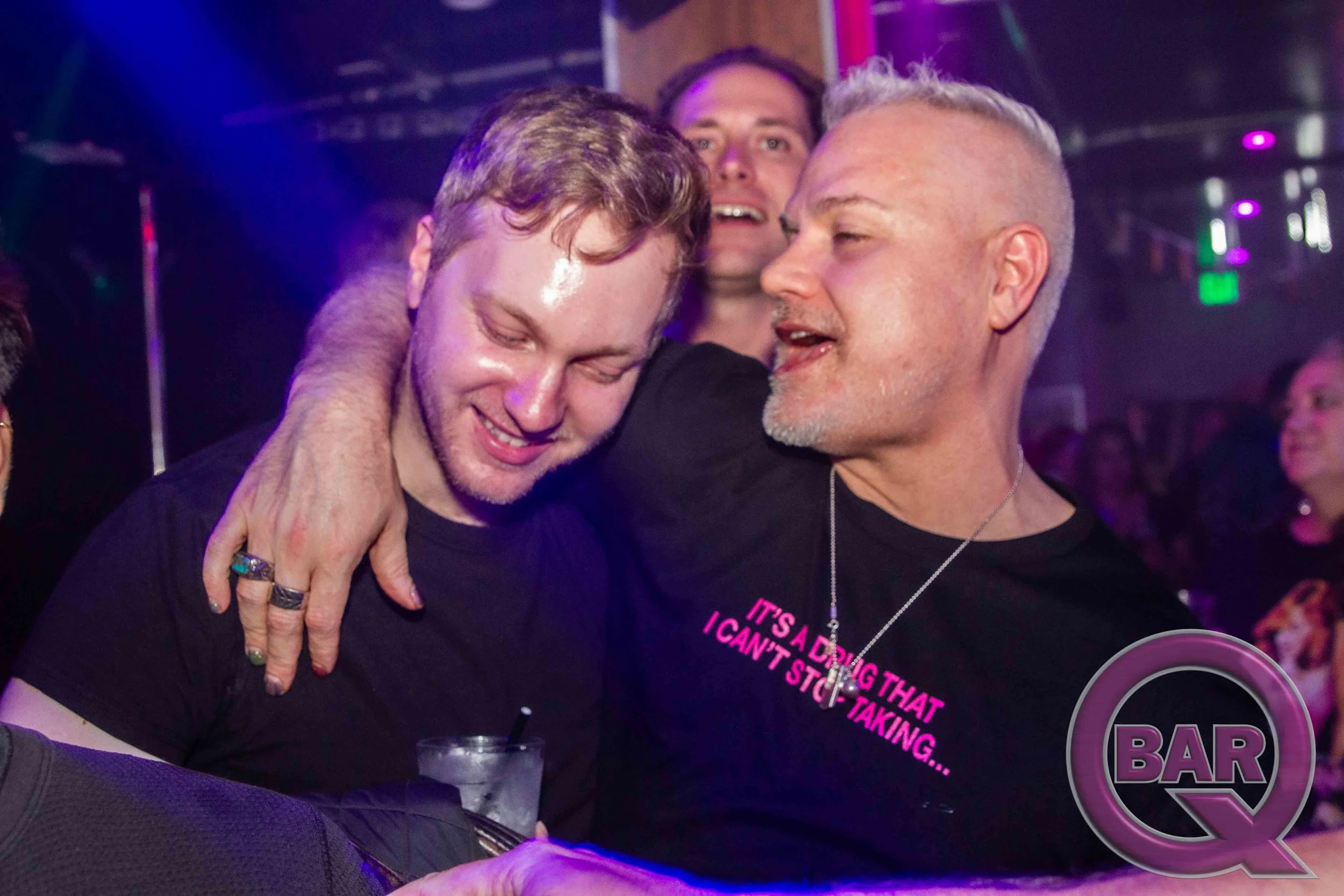 Three men are sharing a close, joyful moment at a bar, with two of them leaning on each other and smiling.