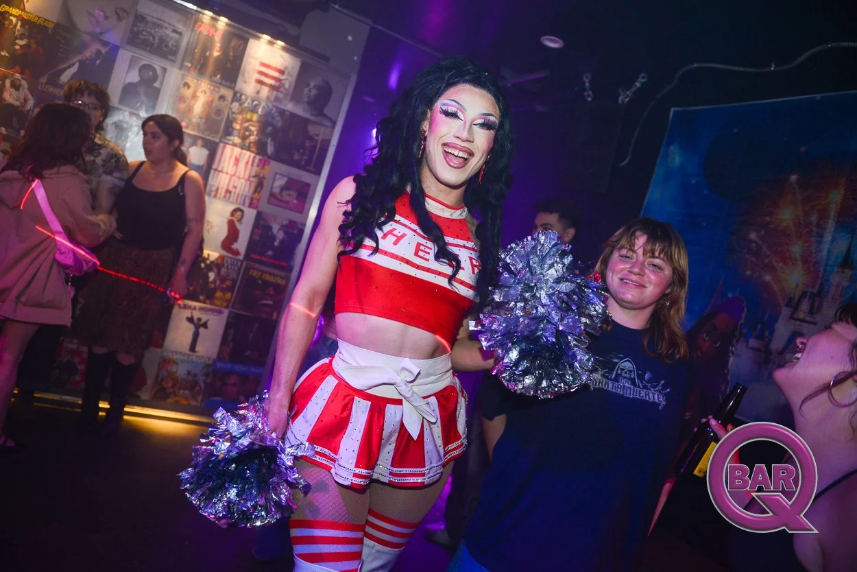 A person dressed as a cheerleader wearing a red and white outfit and holding metallic pom-poms, smiling in a nightclub. Other people are in the background, and the walls are decorated with posters and wall art.