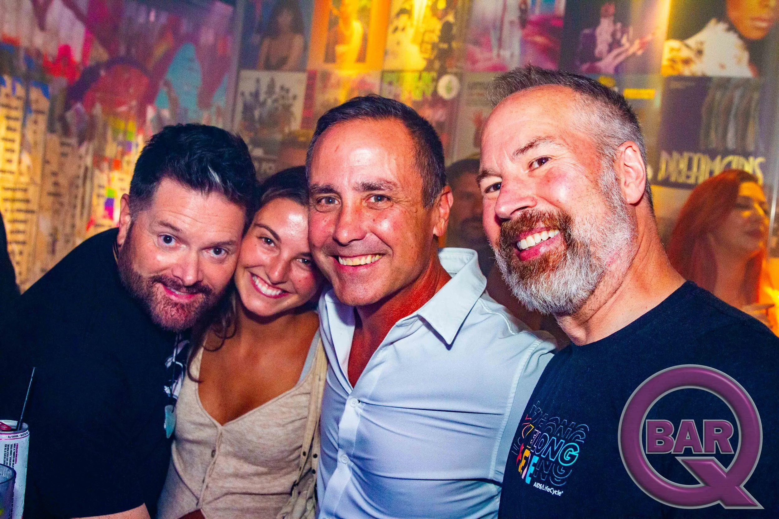 Four smiling people, two men and two women, are close together in a bar or nightclub setting. The background features colorful posters and artwork on the wall.