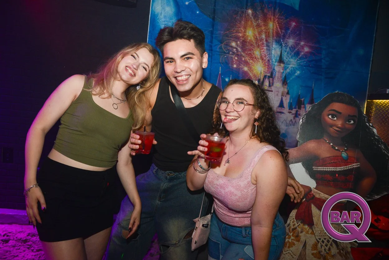 Group of three young adults at a bar, smiling and holding drinks, with a Disney-themed backdrop featuring Moana and a castle with fireworks.