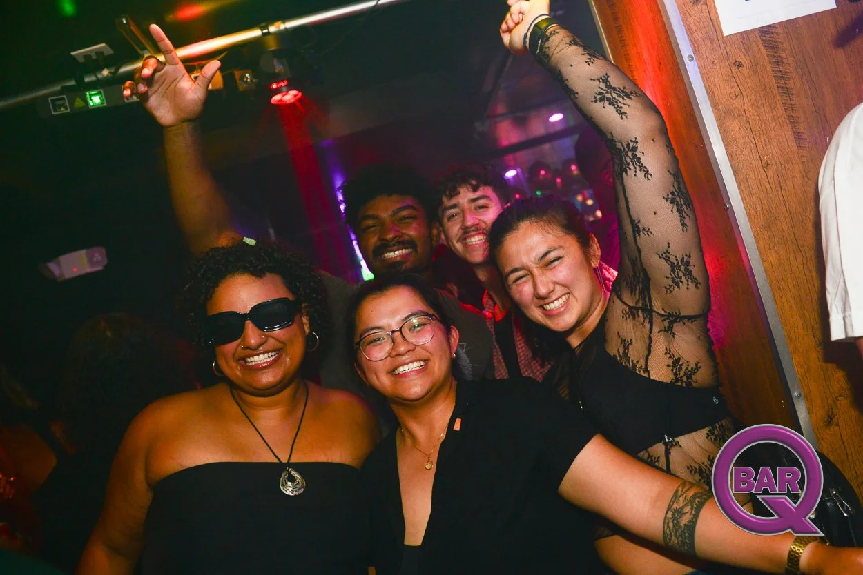 Group of five friends smiling and having fun in a bar or nightclub with colorful lighting, one woman wearing glasses and a black strapless top, another woman with glasses and a black blouse, a woman in a black lace dress with her arm raised, and two 