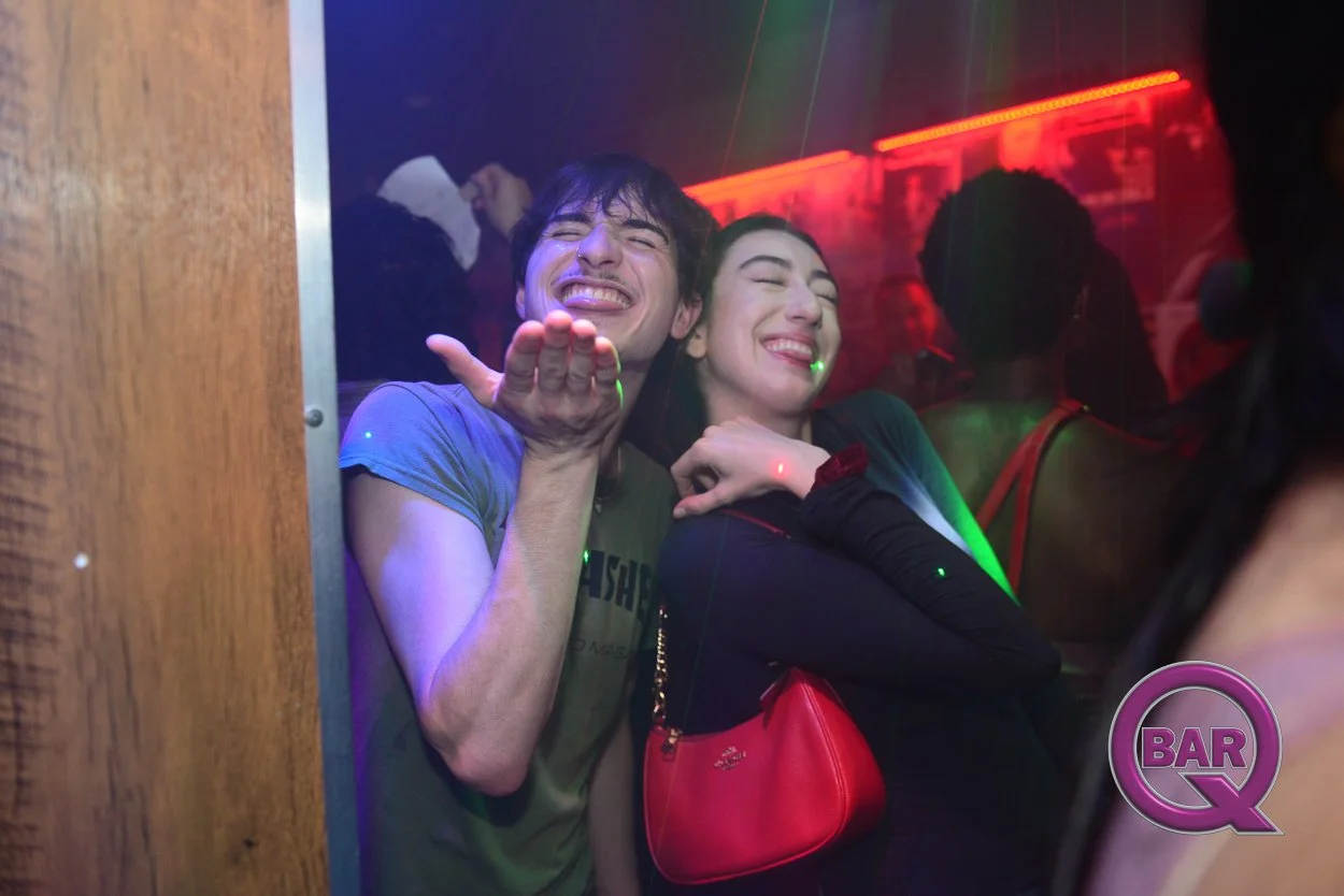 Two young people enjoy dancing at a nightclub with colorful lights, one man playfully blowing a kiss and a woman smiling with a red purse.