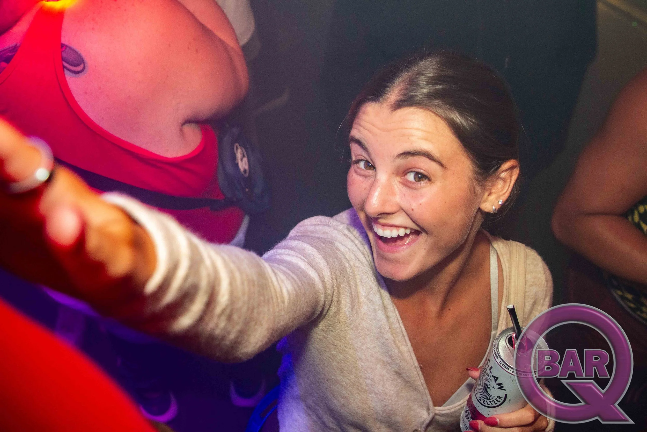 A woman with brown hair smiling while taking a selfie at a bar, holding a can of sparkling water, surrounded by colorful lights.