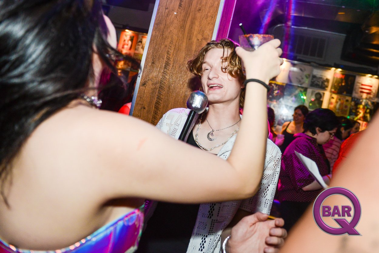 A woman is singing into a microphone at a bar, while another woman is holding up a drink. People are dancing and socializing in the background at a lively nightclub with colorful lighting.