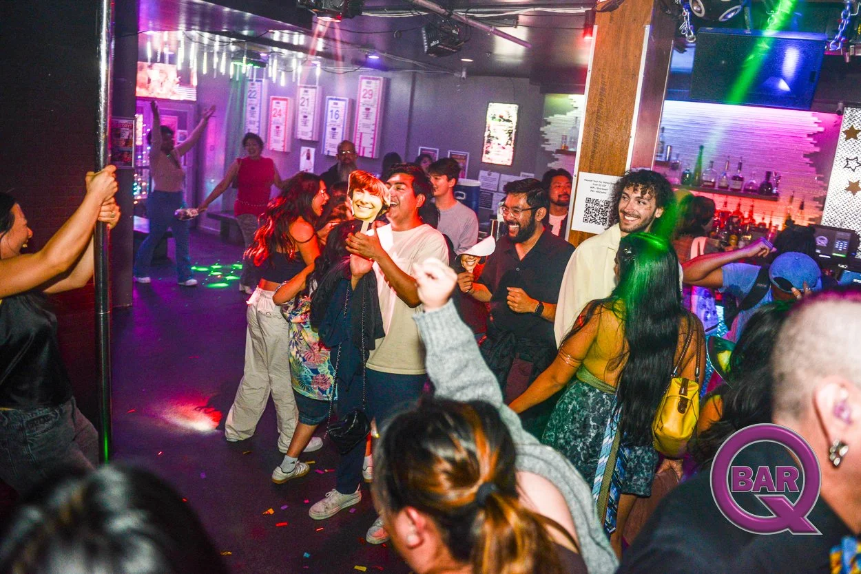 People dancing and socializing at a lively bar or nightclub, with colorful lighting and a bar area in the background.