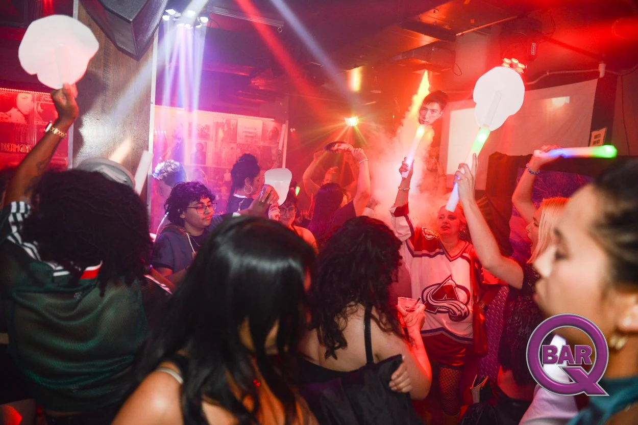 People dancing and enjoying music at a nightclub with laser lights and foam sticks, with a woman wearing a Colorado Avalanche hockey jersey in the foreground and a logo for Bar Q in the bottom right corner.