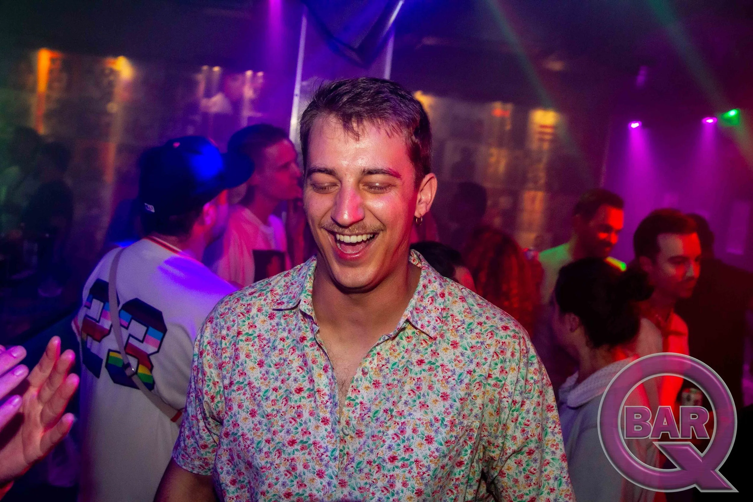 A young man with short brown hair wearing a colorful floral shirt smiling in a lively nightclub with vibrant purple, green, and orange lights and a people dancing in the background.