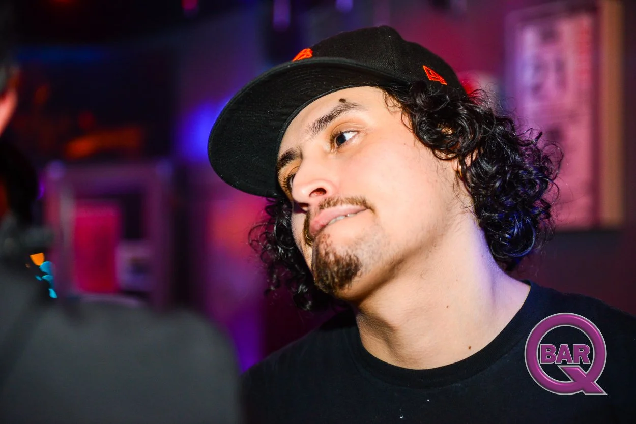 A young man with curly hair, a goatee, and a mustache, wearing a black cap and a black shirt, in a bar or club setting with colorful lighting.