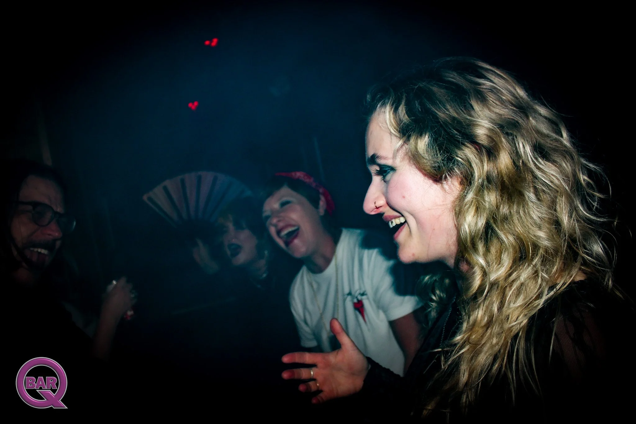 A group of people, including a woman with curly blonde hair and a nose ring, smiling and enjoying themselves at a nightclub or bar with a dark background and neon signs.