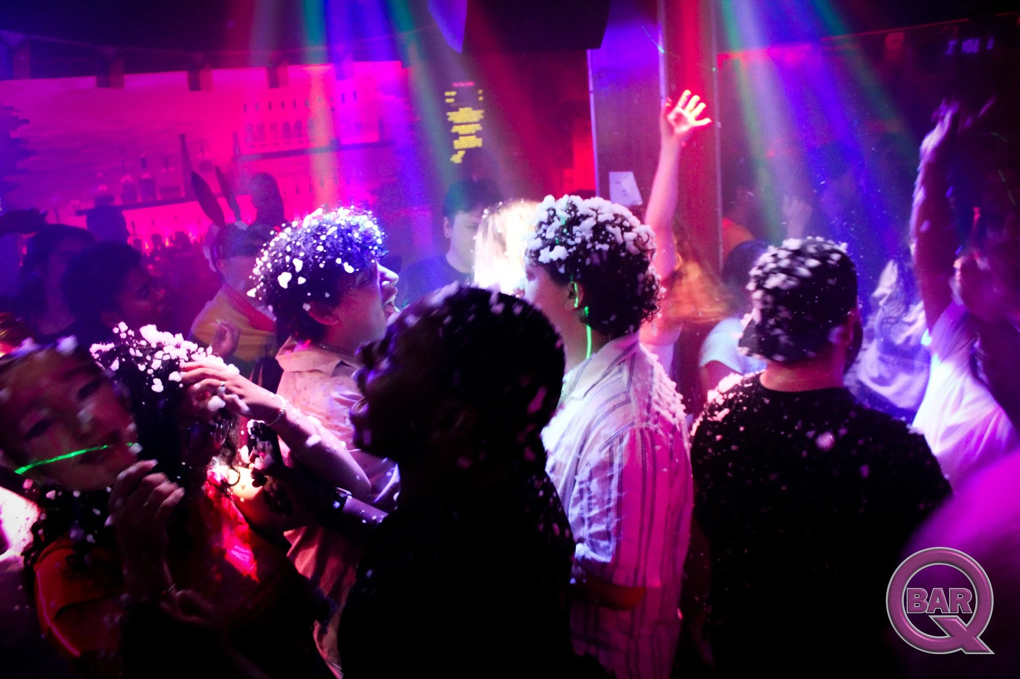 People in a nightclub dancing and socializing under colorful, neon lighting with foam on their hair and shoulders.