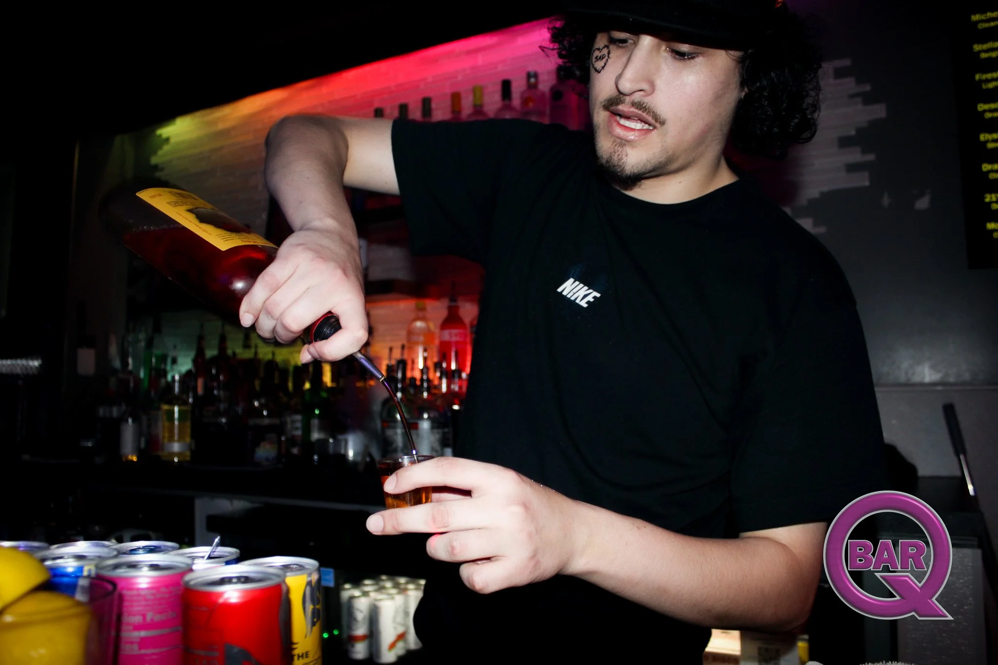 A man wearing a black T-shirt and a black cap pouring a drink from a bottle into a glass at a bar with colorful cans on the counter.