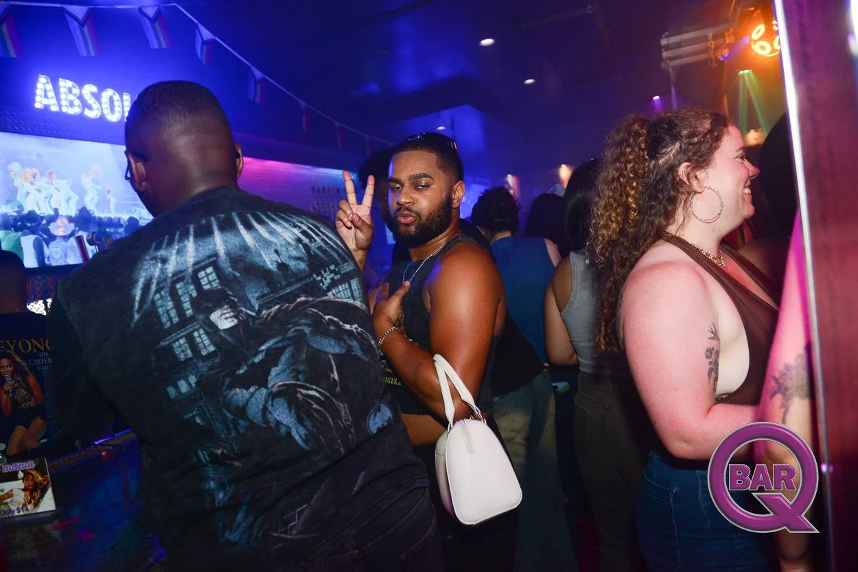 People dancing and socializing at a nightclub with colorful lights and a DJ. A man is making a peace sign and a woman is smiling.