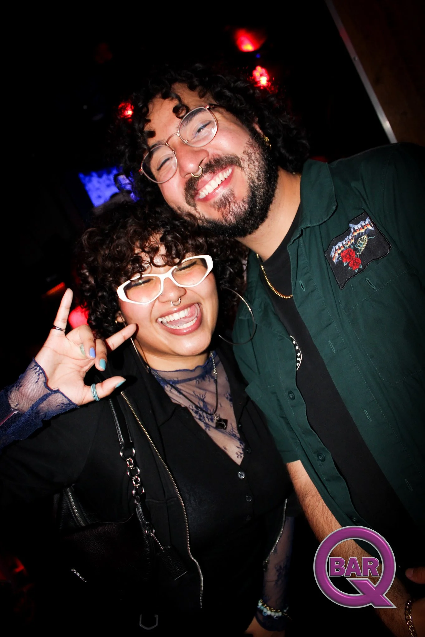 Two smiling individuals, a woman with curly hair, glasses, and septum piercing, and a man with curly hair, glasses, and a beard, posing closely together in a dark setting with red and purple lights, in front of a bar sign.