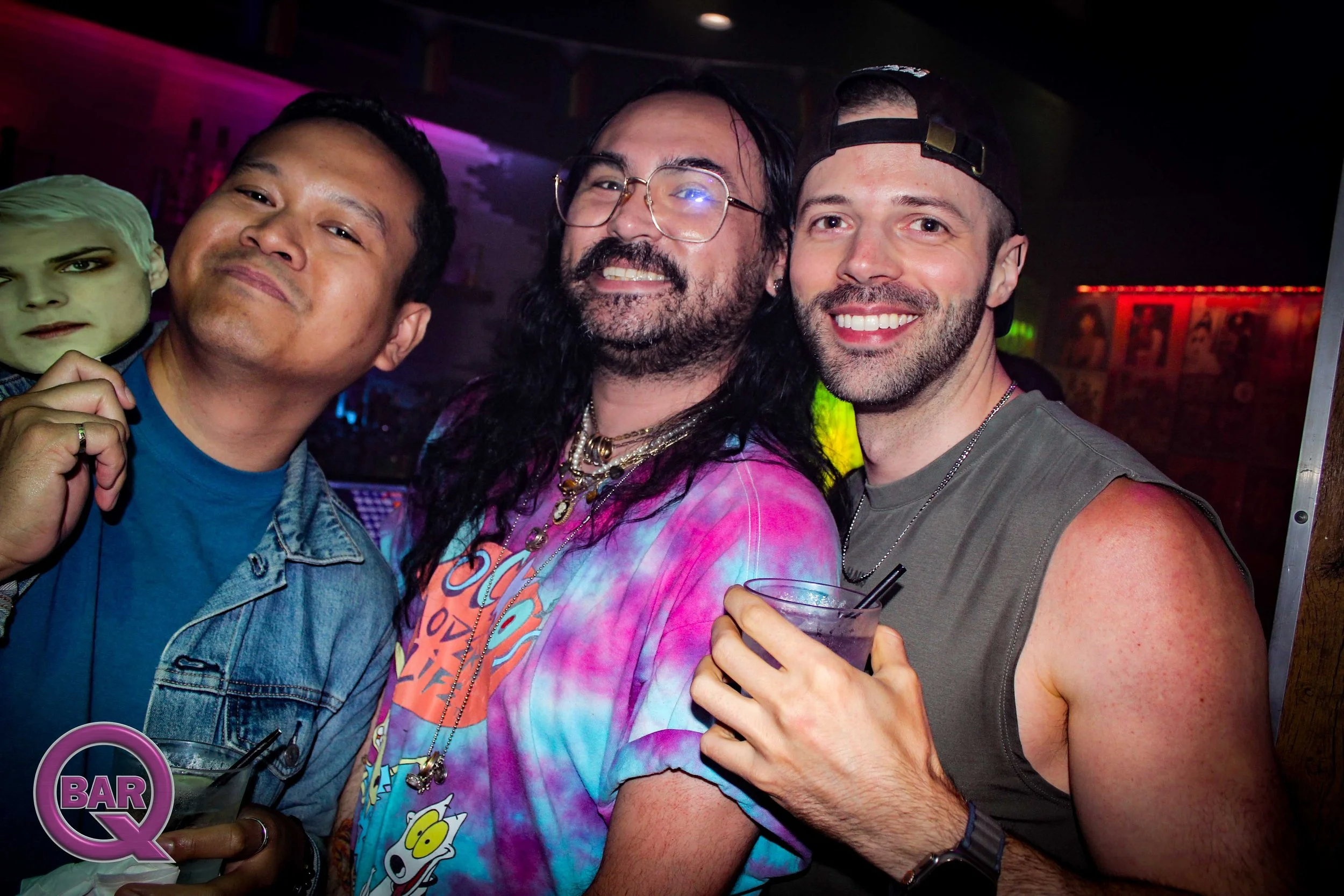 Three men in a bar or nightclub taking a selfie, with colorful lights and decorations in the background.