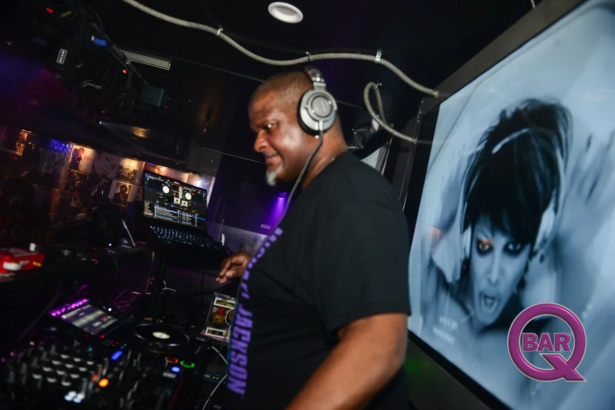 A DJ with brown skin and a beard, wearing headphones and a black T-shirt, is performing in a nightclub. Behind him is a poster featuring a woman with a short, black hairstyle. The DJ equipment, including a laptop and mixer, is visible on the table in