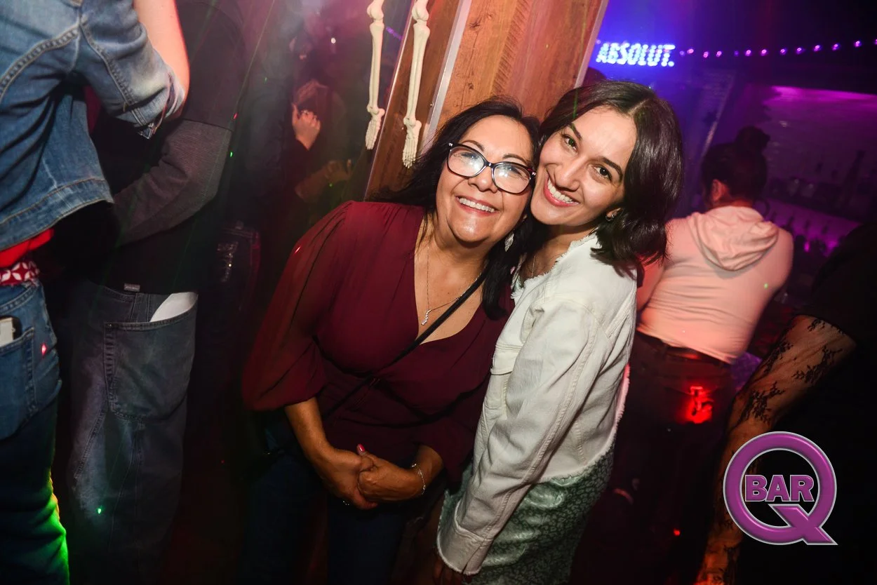 Two women smiling and posing for a photo in a bar or nightclub, with colorful neon lighting in the background.