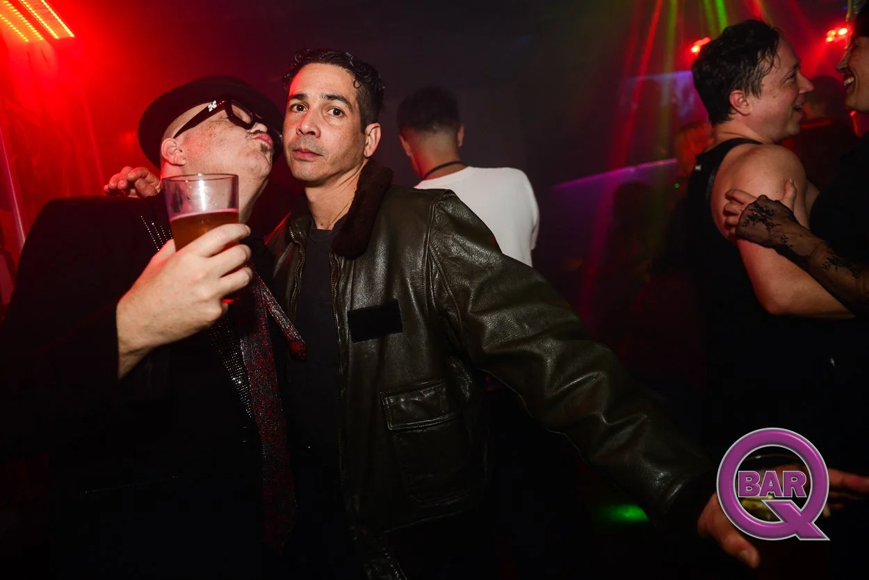 Two men at a nightclub, one holding a drink and leaning in for a kiss, the other looking at the camera. In the background, two women are dancing and smiling. The scene is vibrant with red, purple, and green lights, with a backing sign that says 'BAR 