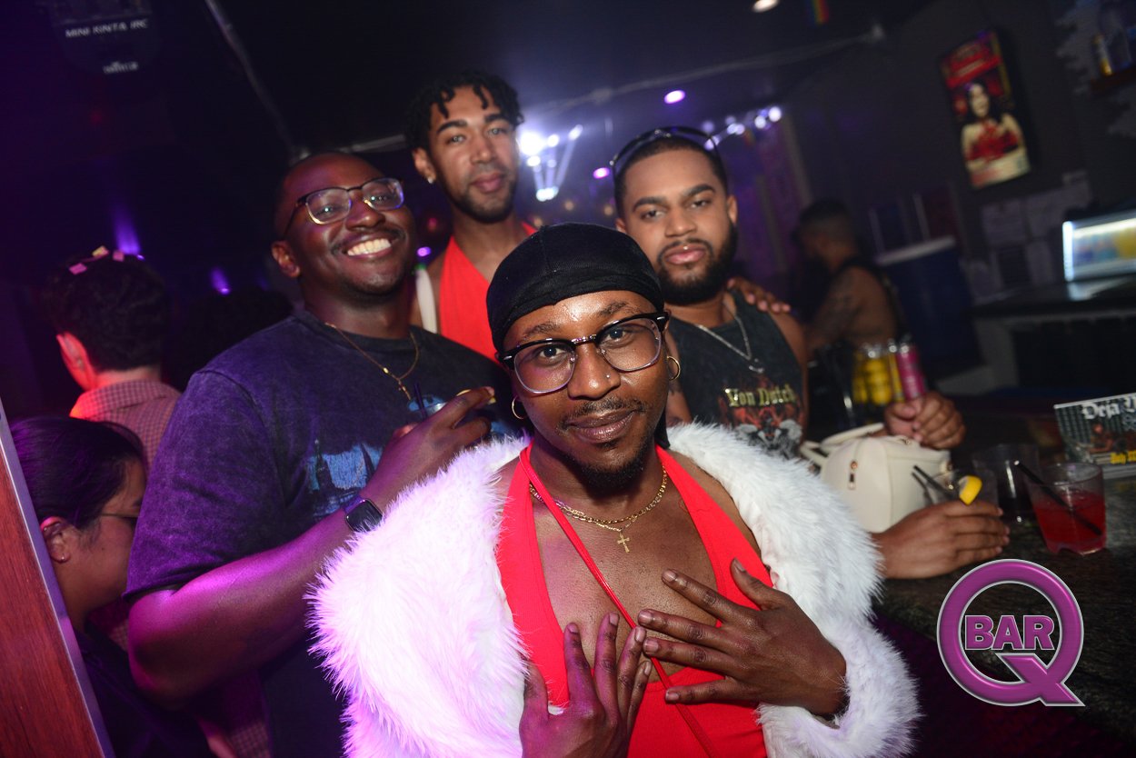 Four men posing together inside a nightclub or bar, with colorful lighting and drinks on the counter. The man in the front is wearing glasses, a black headscarf, a gold necklace, and a white fur coat over a red top, holding his chest with one hand. T
