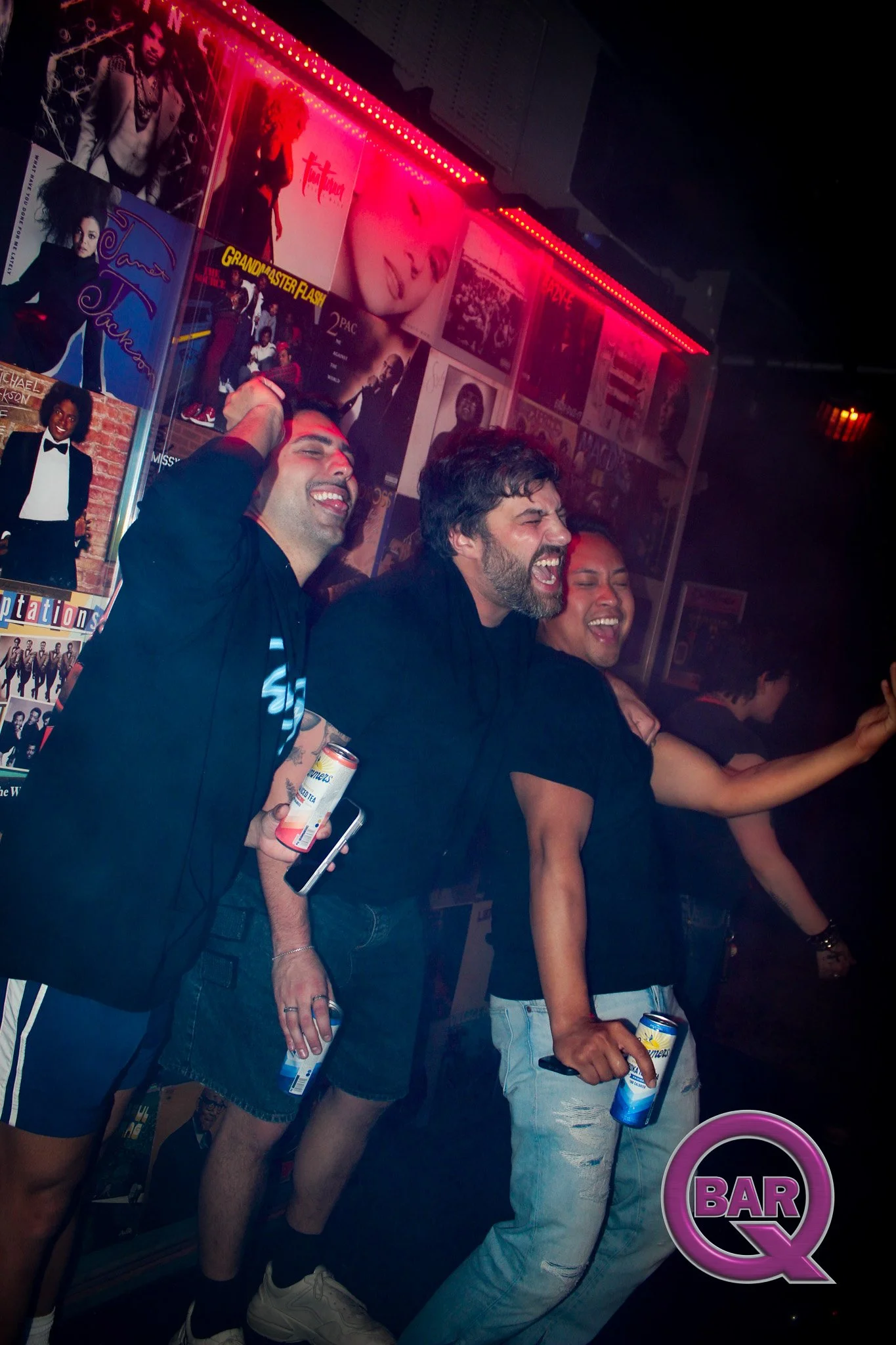 Group of people enjoying themselves at a bar, holding drinks, with colorful posters on the wall and pink neon lighting overhead.