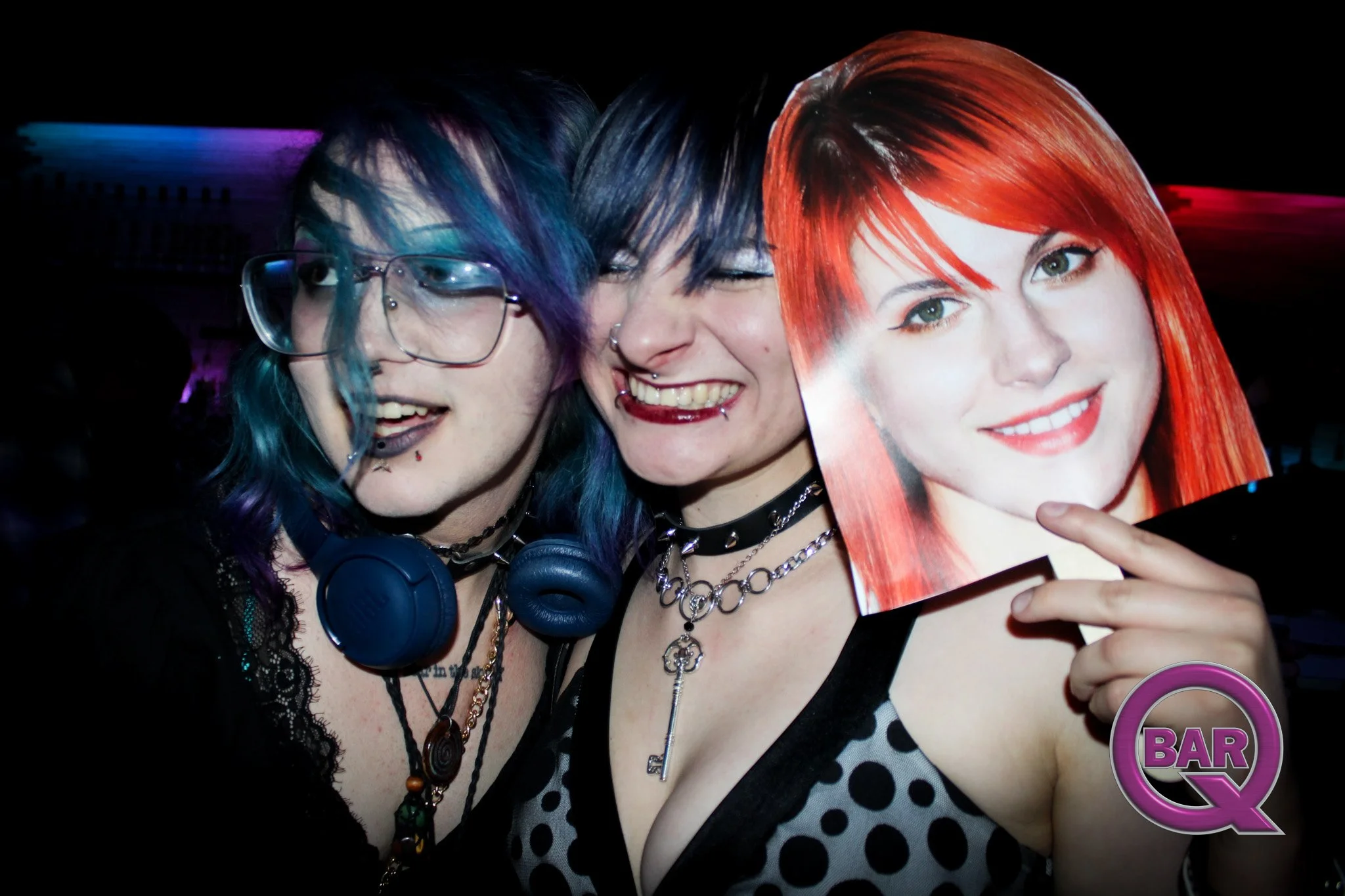Three women at a bar, two with colorful hairstyles and one holding a magazine with a woman's face on the cover.