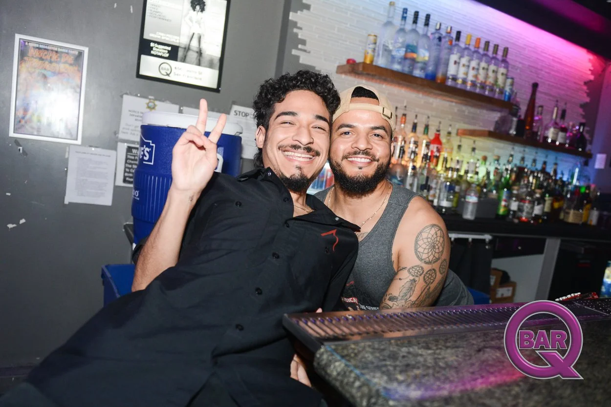 Two smiling men sitting at a bar, one wearing a black shirt and the other in a gray tank top with tattoos, posing happily for the camera with one making a peace sign. The bar area has liquor bottles on shelves and colorful lighting.
