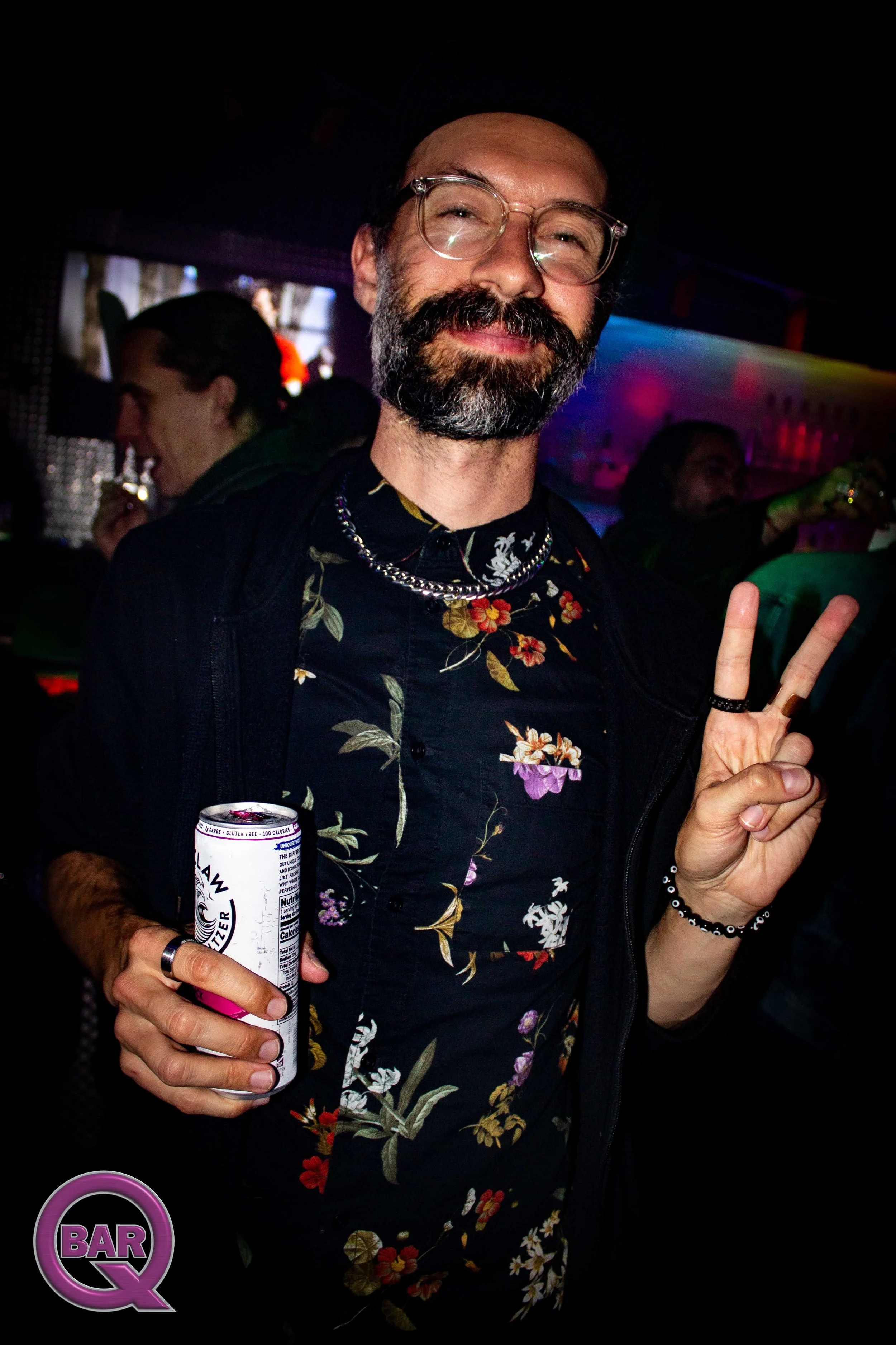 A man with glasses and a beard, smiling and making a peace sign with his fingers, holding a canned beverage. He's wearing a floral shirt and a beaded bracelet, in a dimly lit bar with other people in the background.