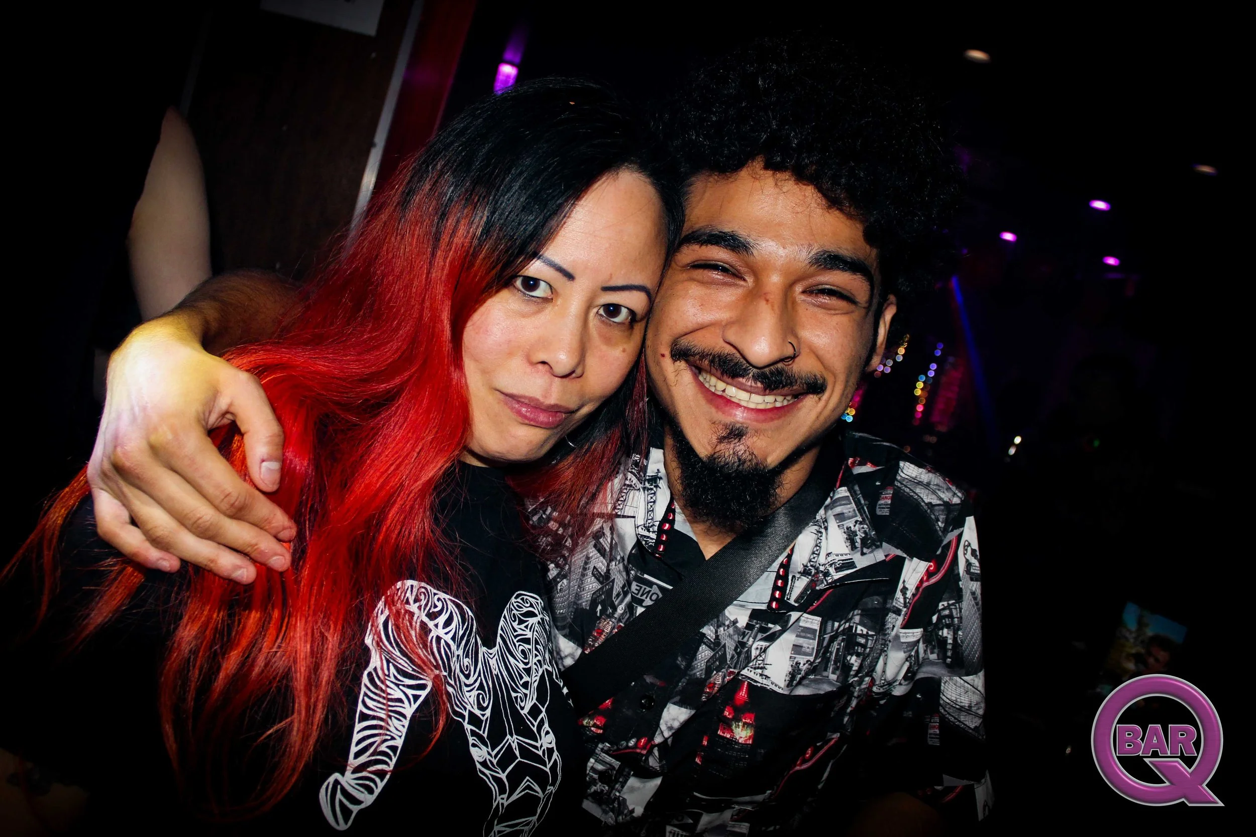 Two friends hugging at a bar, one with black and red hair and the other with curly hair and a nose ring, smiling in a dimly lit venue with purple lighting.