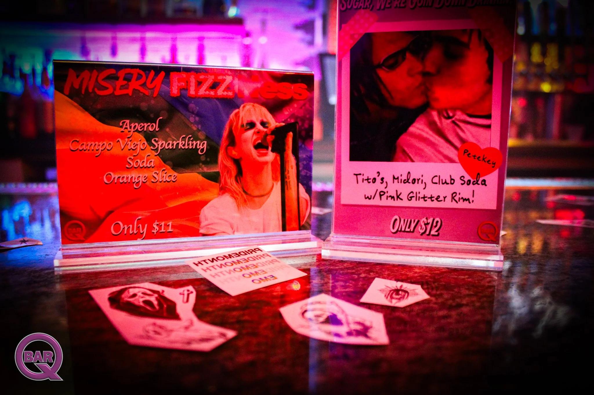 Two colorful posters on a bar counter illuminated by neon lights. The left poster features a woman singing into a microphone with the words 'Miserly Fizzness' and list of drinks, priced at $11. The right poster shows a person with glasses sipping fro