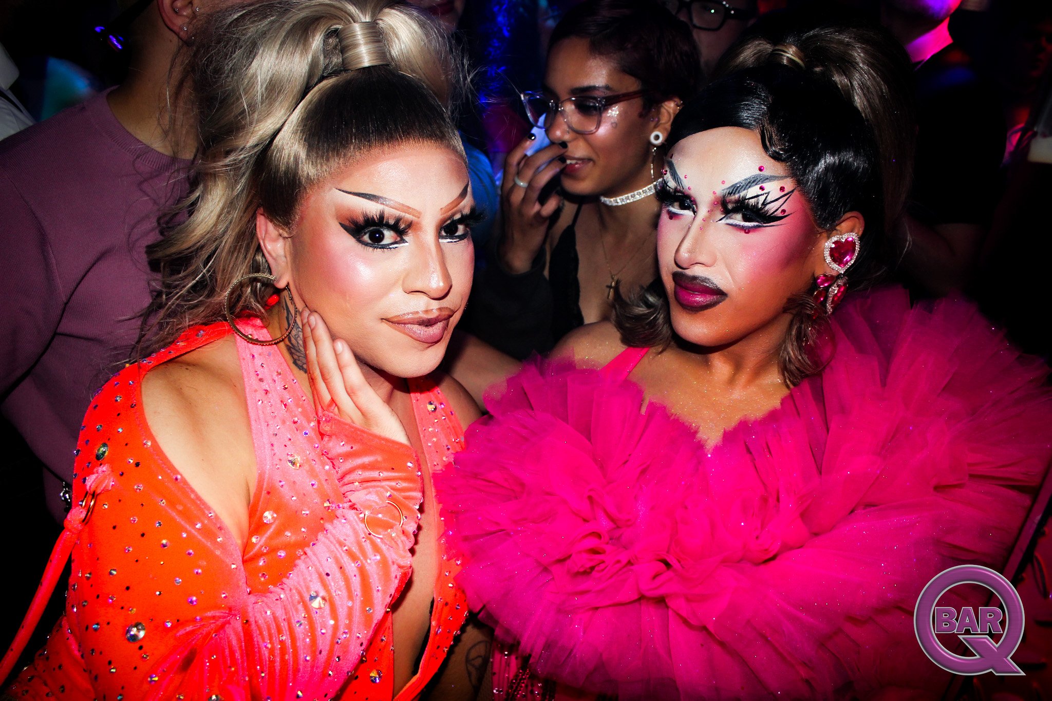 Two women with elaborate makeup and colorful outfits at a crowded event. One in pink with big earrings and the other in bright orange with decorated sleeves. The background has other people and colorful lighting.